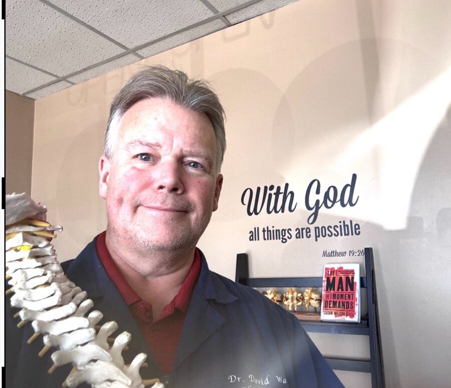 Dr. David Waring, DC in his Sterling Heights clinic holding a spine model, beneath his 'With God all things are possible' sign